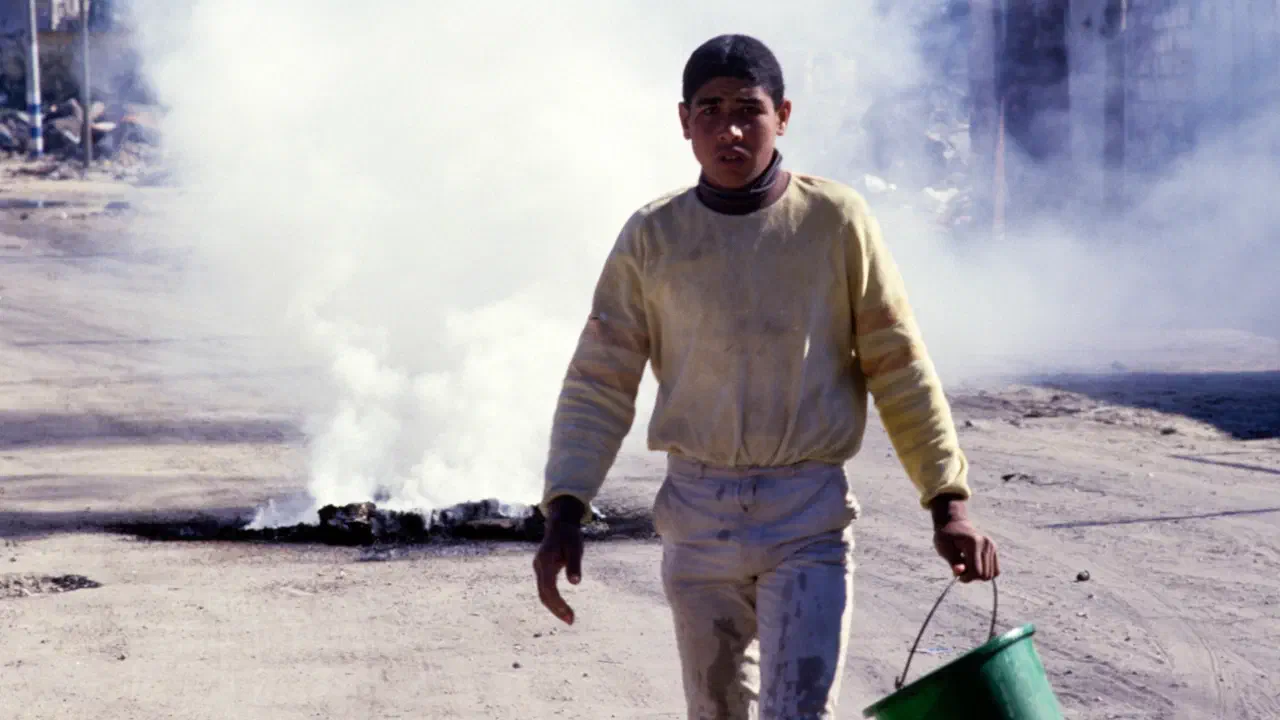 A Palestinian boy carries a bucket while a pile of ash smoulders on the road behind him.